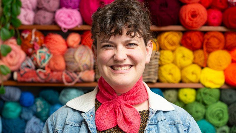 Person smiling in front of colorful yarn displayed on shelves, wearing a denim jacket and a red knitted scarf.