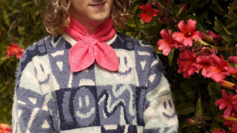 Person with long curly hair in a smiley face sweater and red scarf, near vibrant pink flowers.