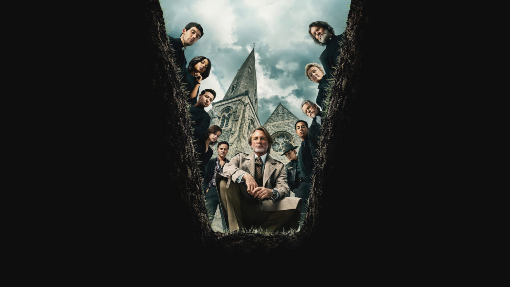 A group of people in dark clothing and one man in beige look down into a grave with a church in the background.