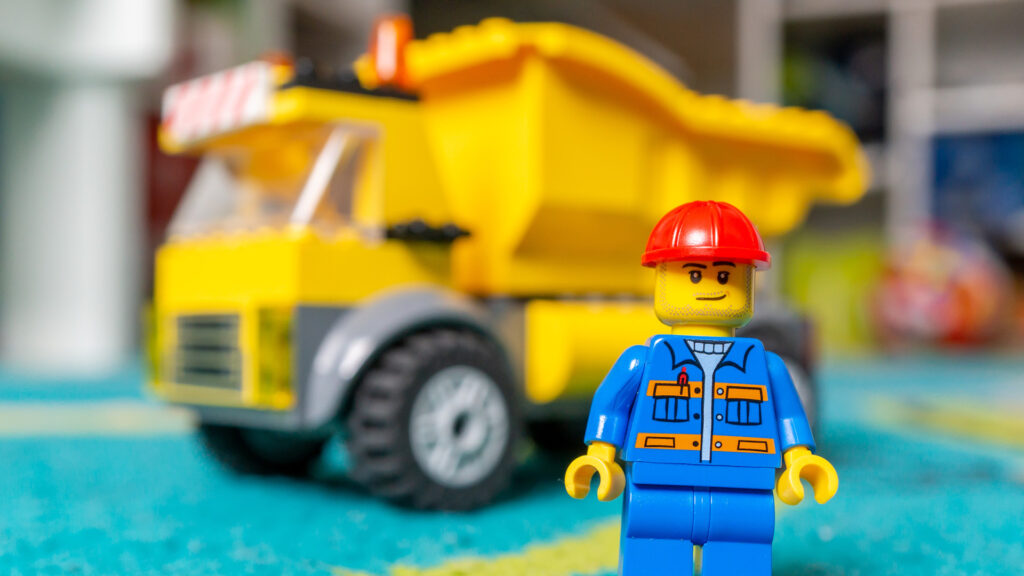 Close-up of a LEGO construction worker figurine with a red helmet and blue uniform, standing in front of a yellow dump truck.