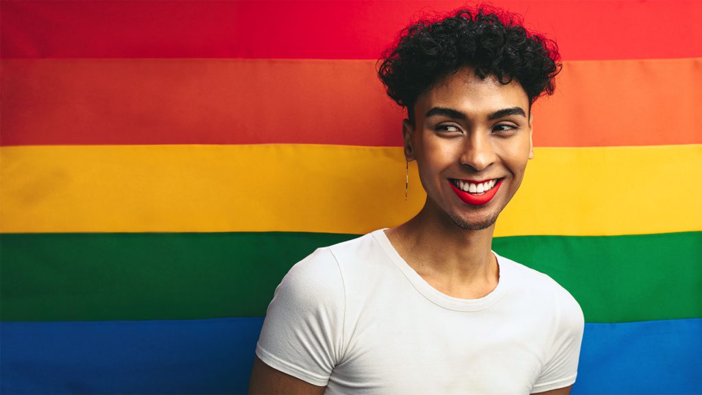 A person with curly hair and red lipstick smiles in front of a rainbow flag, wearing a white shirt.