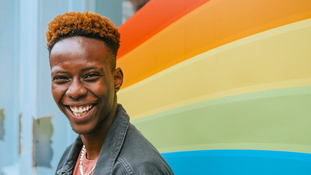 Smiling person with short curly hair stands in front of a colorful wall painted with a rainbow.