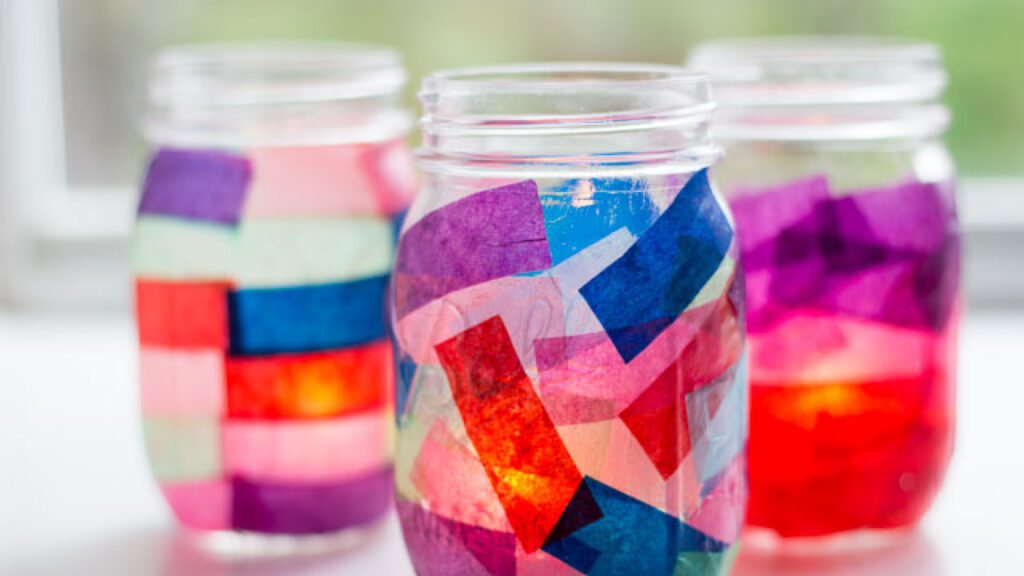 Three mason jars decorated with colorful tissue paper, illuminated from inside, sitting on a white surface.