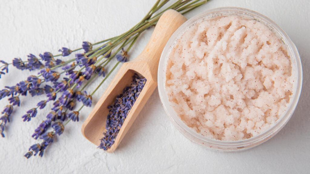 A jar of lavender body scrub, a wooden scoop, and fresh lavender sprigs on a white surface.