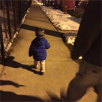 A small child in a blue jacket walks down a sidewalk at night, accompanied by an adult. Snow is seen on the ground.