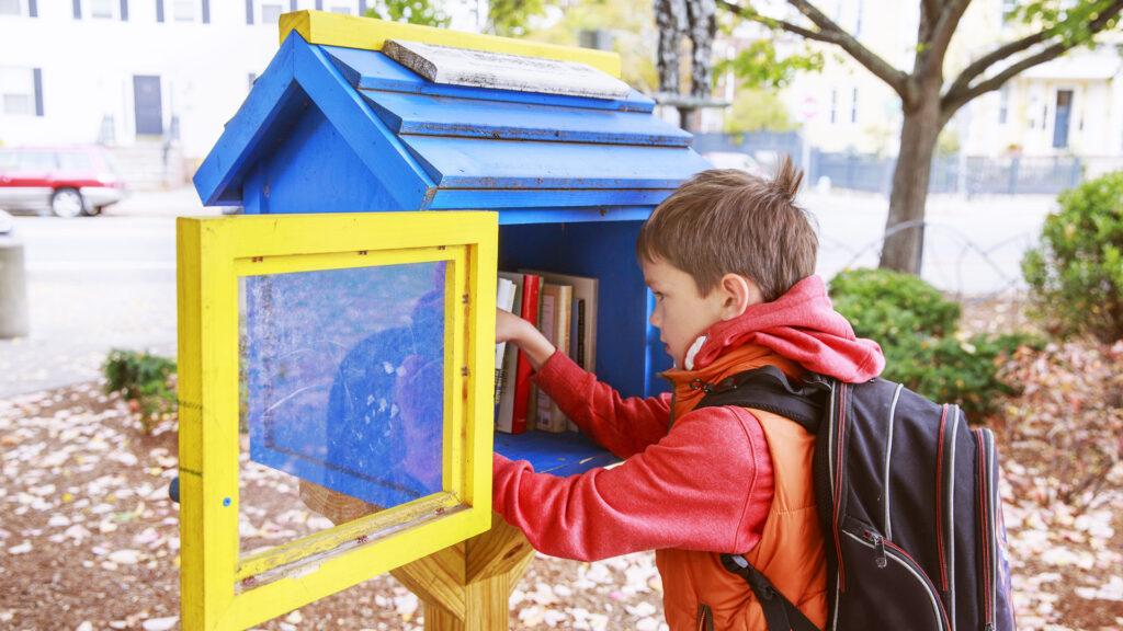 A child with a backpack selects a book from a small blue and yellow outdoor library.