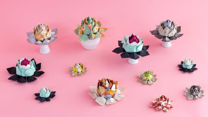 Various origami flowers in vibrant colors on pink background, some displayed in small white vases.
