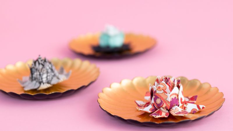 Three origami flowers on gold-rimmed plates are displayed against a pink background.