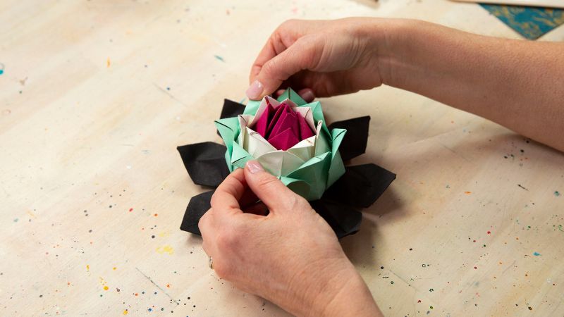 Hands folding a colorful paper flower with black, green, white, and pink petals on a wooden table.