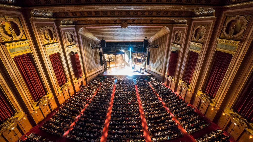 An ornate theater with red seats and walls is filled with a large audience watching a stage performance.