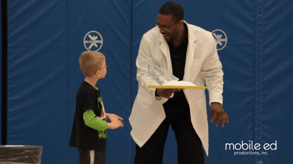 Man in lab coat showing ice to a boy in a gymnasium with blue padded walls.