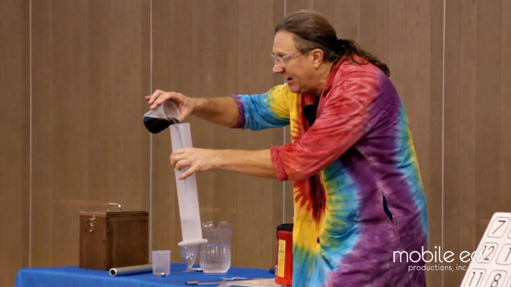 Person in a colorful lab coat pouring liquid during a science demonstration on stage with props and equipment.