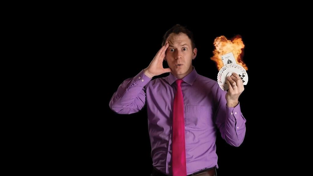 A surprised man in a purple shirt holds flaming playing cards against a black background.