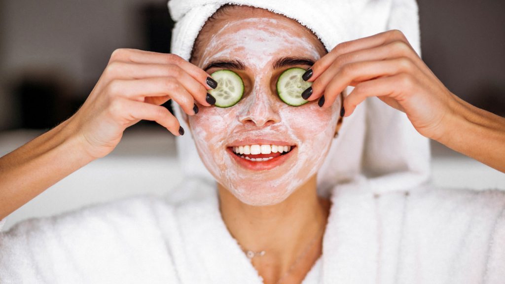 A person wearing a white towel turban and face mask, holding cucumber slices over their eyes and smiling.