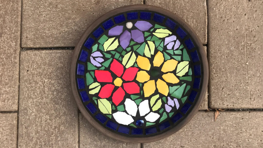 Round mosaic stepping stone with colorful flower patterns on a brown brick patio.