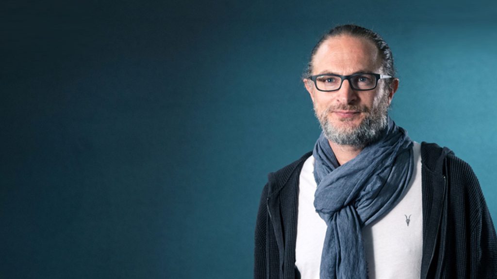 A man with glasses, a beard, and long hair stands against a blue background, wearing a white shirt, black jacket, and scarf.