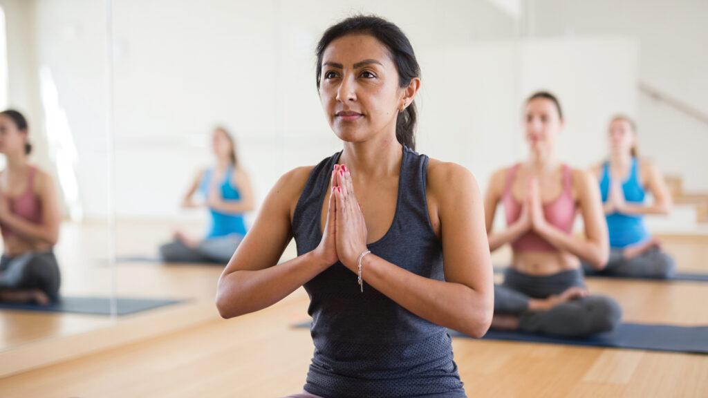 Woman in workout clothes sitting cross-legged in a yoga class, hands in prayer pose, with others in the background.