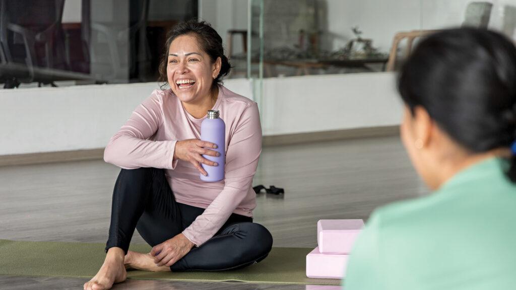 Smiling woman sits on a yoga mat holding a water bottle, talking to another person in a studio.
