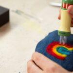 Hands using a felting tool to create a rainbow pattern on blue fabric.