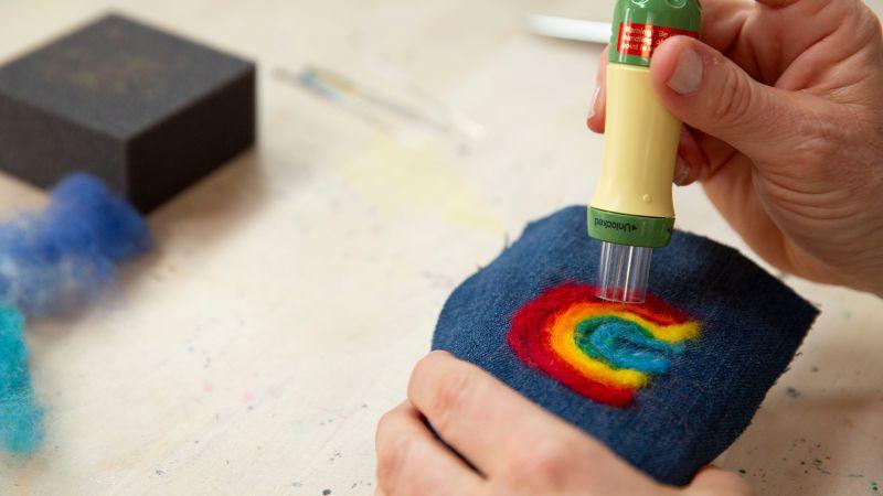 Hands using a felting tool to create a rainbow pattern on blue fabric.