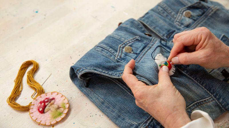 Hands embroidering a design on blue jeans, with embroidery floss and a mushroom patch nearby.