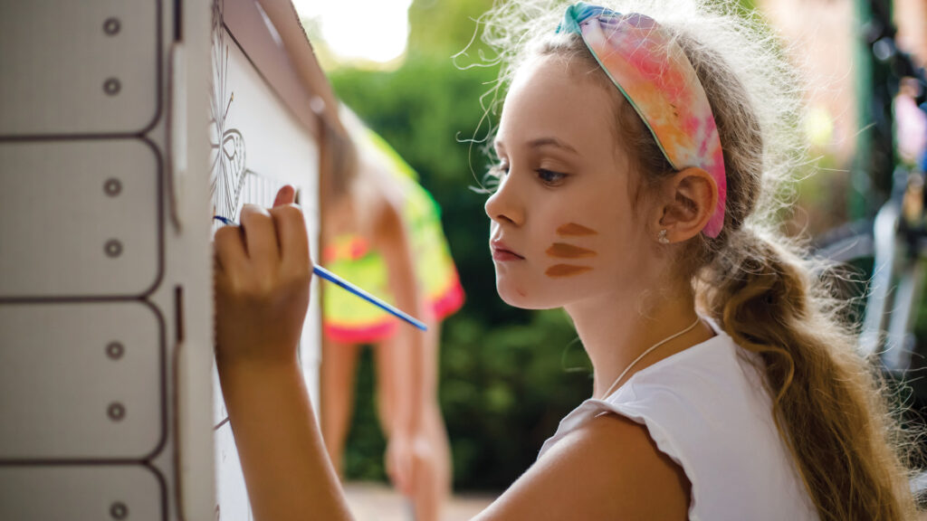 Young girl painting on a canvas outdoors, with a focused expression and colorful headband. Face has brown paint streaks.