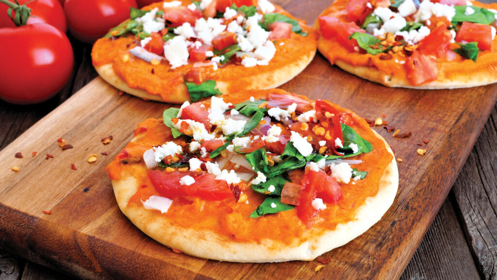 Mini pizzas topped with tomatoes, spinach, onions, feta cheese, and red pepper flakes on a wooden board.