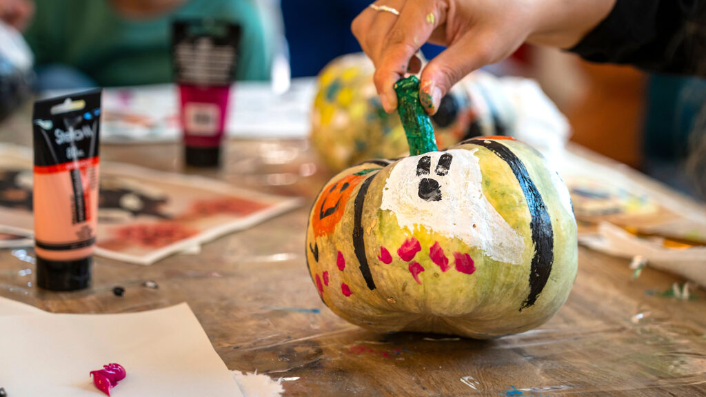 A hand paints a ghost face on a pumpkin. Art supplies and other painted pumpkins are on the table.
