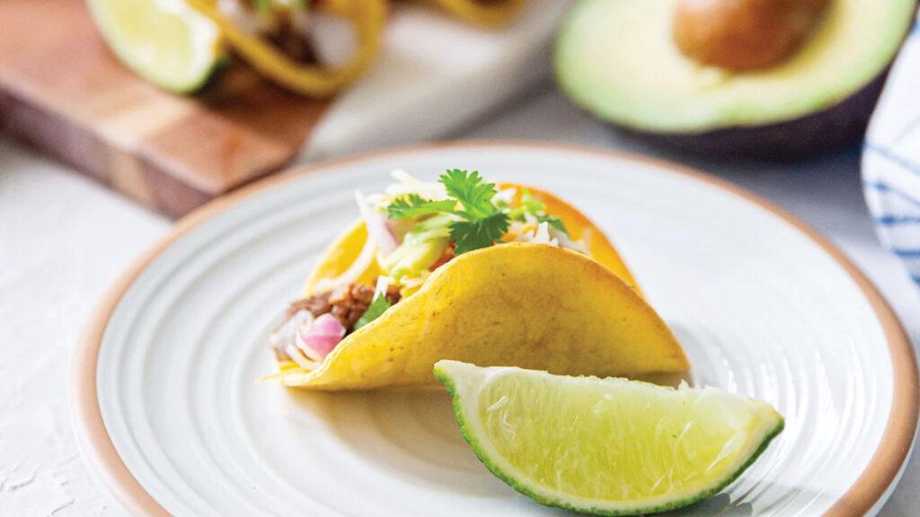 A taco with cilantro and onions on a plate with a lime wedge, and half an avocado in the background.