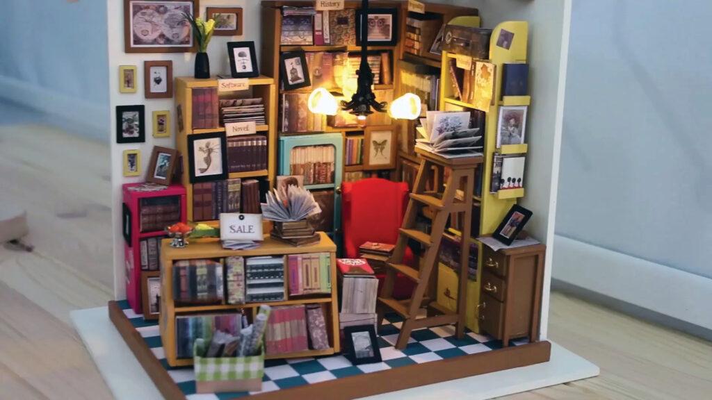 Miniature cozy bookstore scene with bookshelves, framed pictures, a red chair, ladder, and warm lighting.