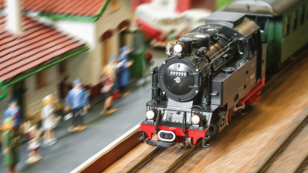 A model steam train on tracks passes a miniature station with blurred figures waiting nearby.