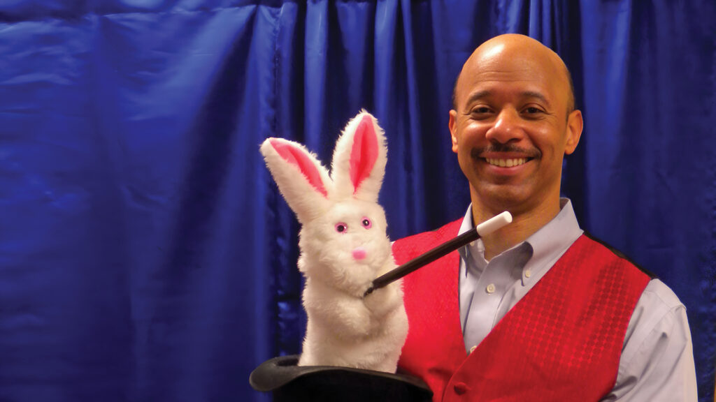 A smiling magician holds a top hat with a white rabbit puppet and magic wand, in front of a blue curtain.
