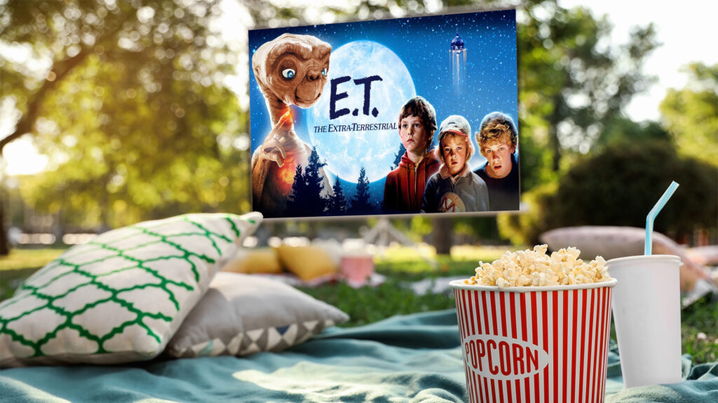 Popcorn and drink on a picnic blanket at an outdoor movie showing "E.T. the Extra-Terrestrial" on a screen.