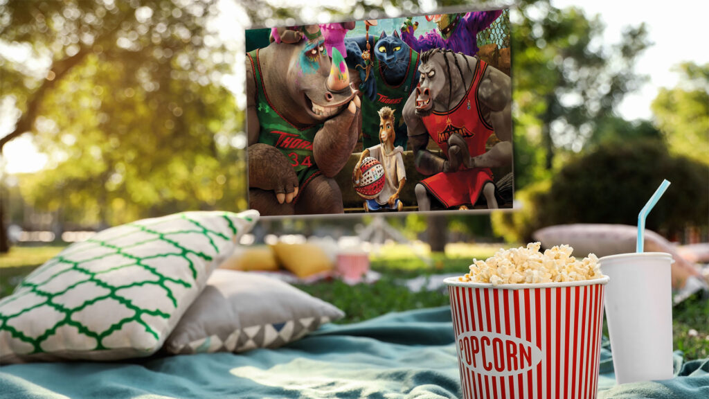 Outdoor movie setup with popcorn, drink, pillows, and an animated film playing on a screen in a sunny park.