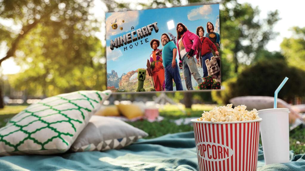 Outdoor movie setup with popcorn, pillows, and "Minecraft" movie poster on screen in a park.