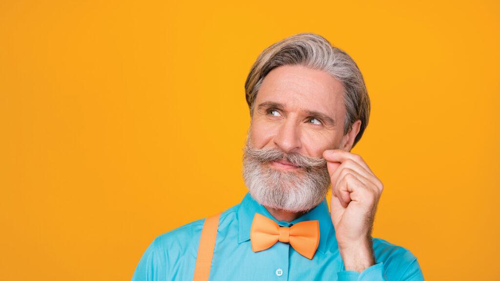 Smiling gray-haired man with a mustache, wearing a blue shirt and orange bow tie, against an orange background.