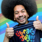 Smiling man with a large afro holding up a "Library Joy!!" shirt, with a rainbow background.