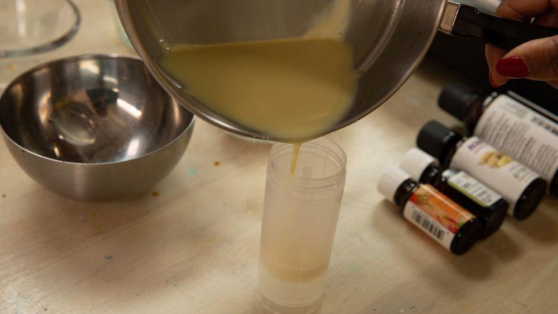 A person pours a creamy mixture from a saucepan into a plastic tube, with bottles and a metal bowl nearby.