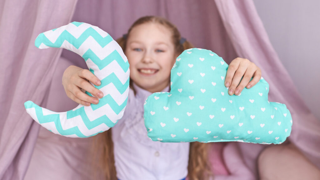 Smiling child holds a crescent moon pillow and a cloud pillow with heart patterns in both hands.