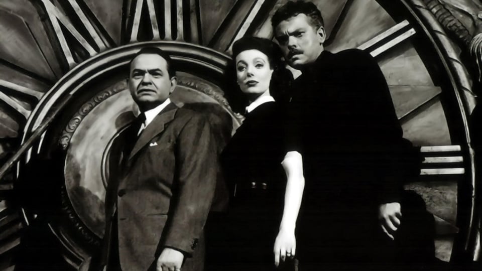 Three people in formal attire standing in front of a large clock face. Black and white photo with dramatic lighting.