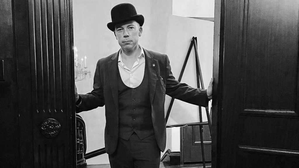 A man in a suit and bowler hat stands in an open doorway, holding the doors with both hands. The image is in black and white.