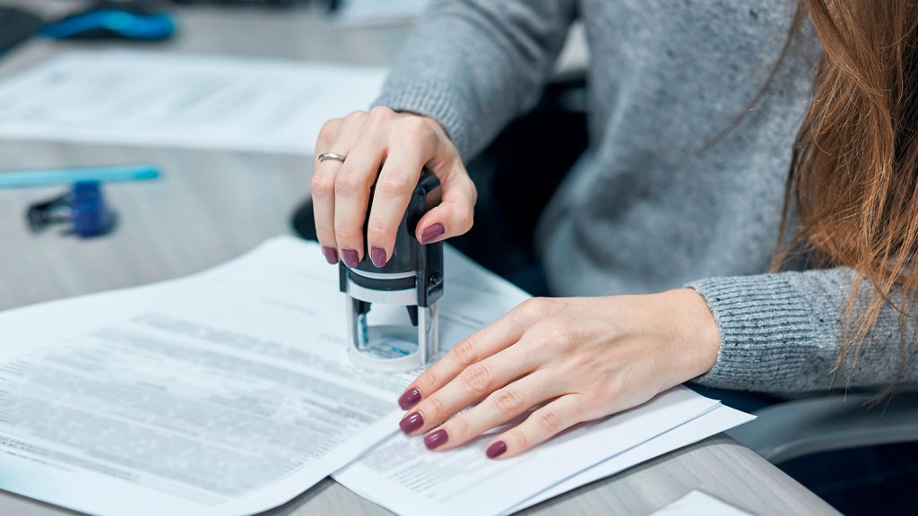 A person stamps a document on a desk while holding the paper steady with their other hand.