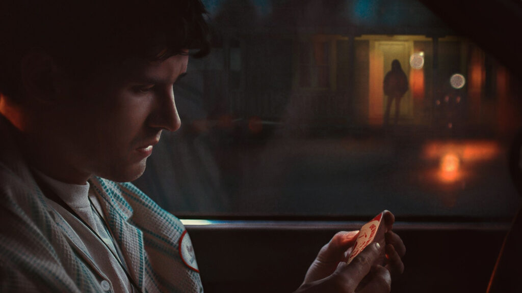 A man sits in a car at night, looking at an object in his hands; a figure stands outside in the background.
