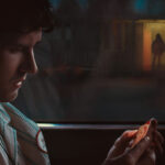 A man sits in a car at night, looking at an object in his hands; a figure stands outside in the background.