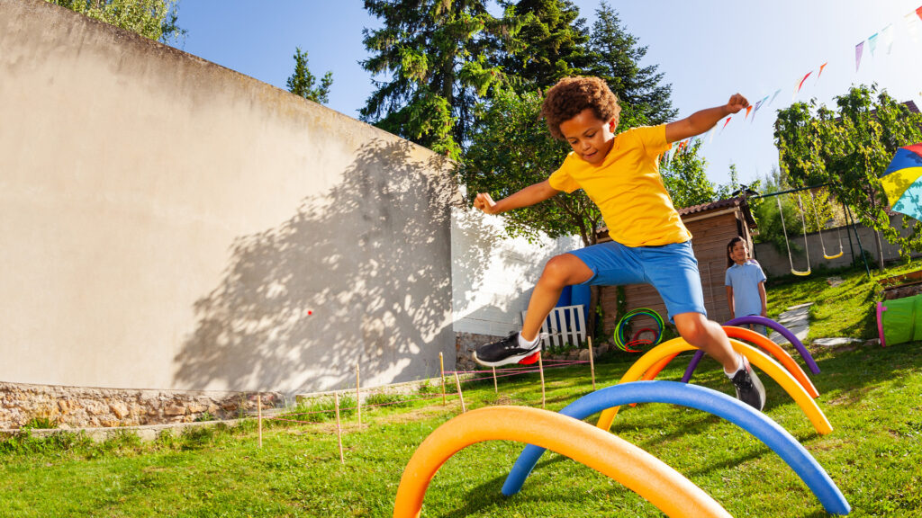 A young boy in a yellow shirt and blue shorts jumps over colorful arches in a sunny backyard playground.