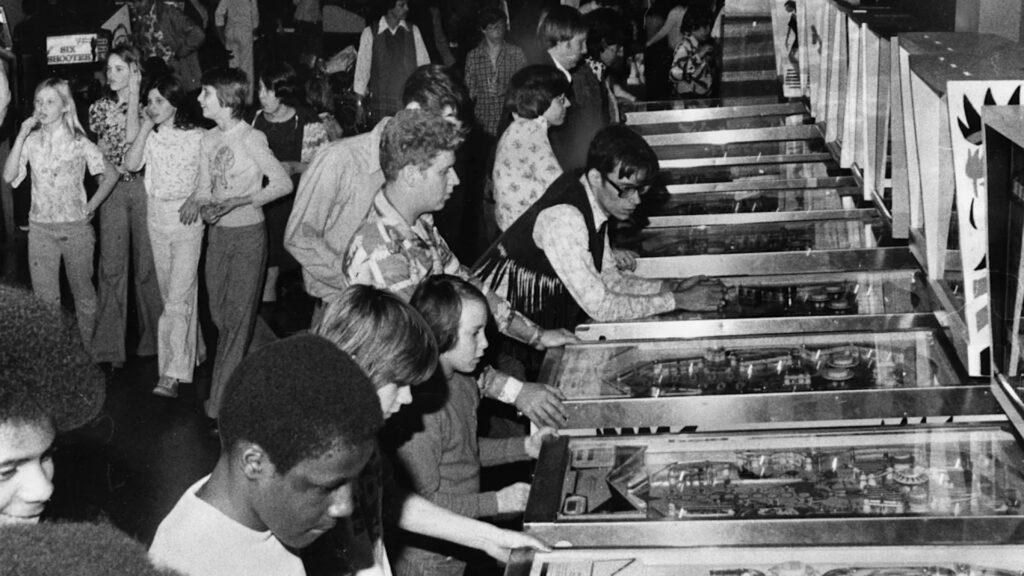 A crowd of people play pinball machines in a busy arcade, likely from the 1970s based on clothing styles.