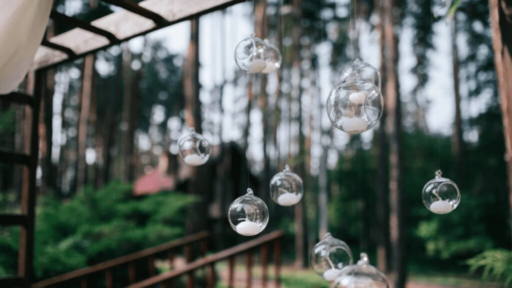 Hanging glass globes with candles inside are set against a background of trees and a wooden structure.