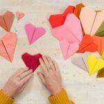 Hands folding a red paper heart among various colorful paper hearts on a table.