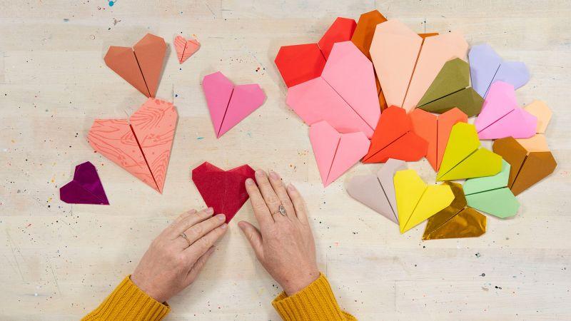 Hands folding a red paper heart among various colorful paper hearts on a table.