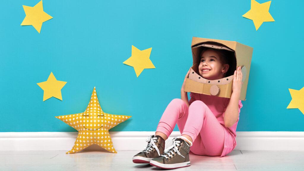 Smiling child sits on floor wearing a cardboard box helmet, surrounded by yellow stars on a blue wall.